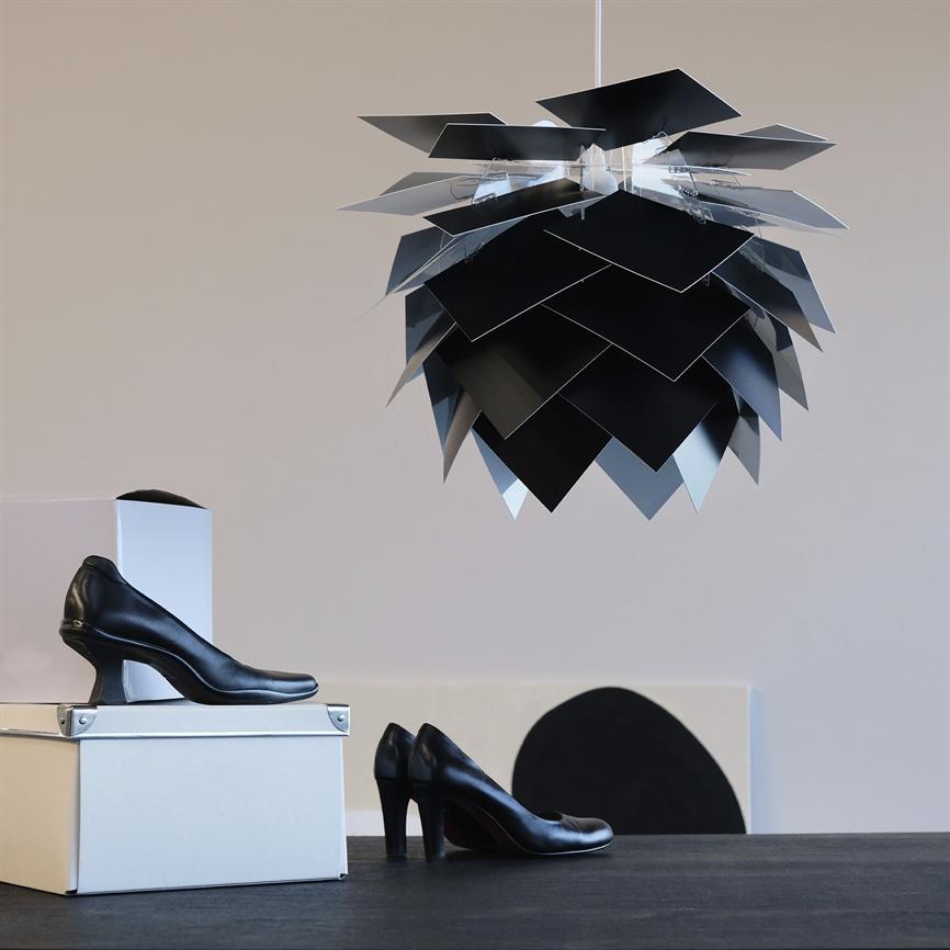Black, geometric pendant lamp hangs from the ceiling like a layered leaf pattern, above a table with black women's shoes placed on white shoe storage boxes in a light, neutral room.