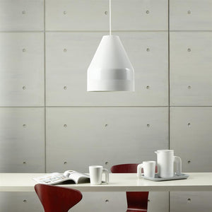 White pendant hangs over a white table; white cups and jug are on a plate, an open magazine is on the table, red chairs and a light gray concrete element wall in the background.