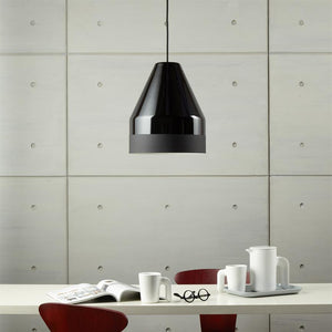 A black, shiny pendant hangs over a table with open magazine, glasses, white mugs and pitcher; the setting is a minimalist dining room with square concrete panels and red chairs.