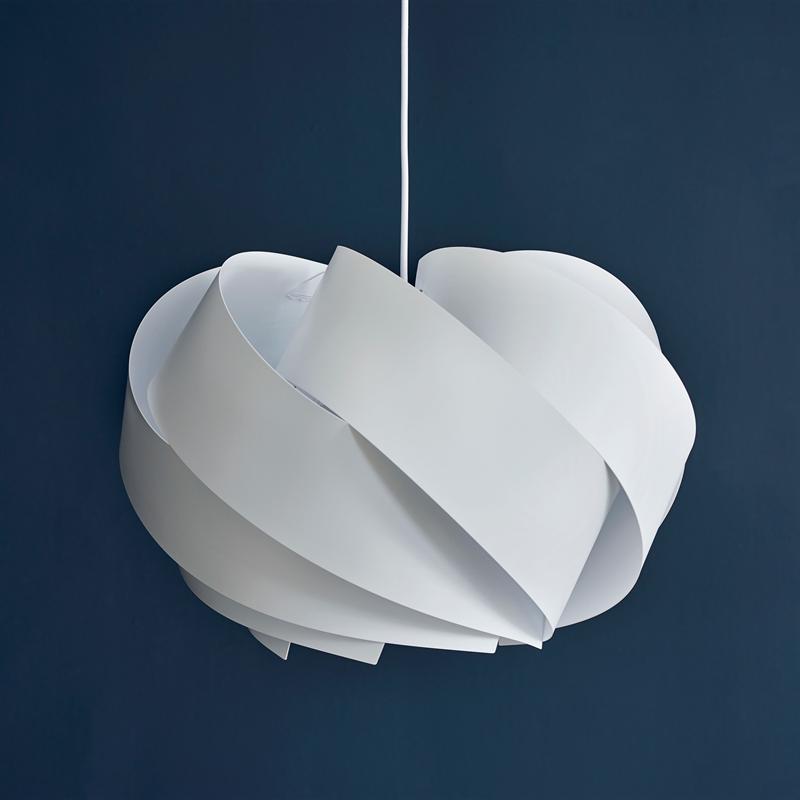 White lampshade hangs from a thin wire; overlapping spiral plates form a soft, sculptural form, with a uniform dark blue wall as a background.