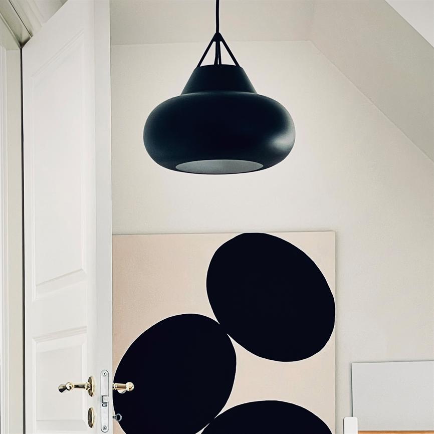 Black hanging lamp hovers above a canvas with three large black circles, placed against a white wall in a bright room; a partially open white door with a brass handle in the foreground.