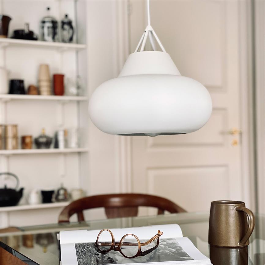 White ceiling lamp hangs above a glass table; on the table is an open book with glasses and a brown mug; shelves with china and a chair in the background.