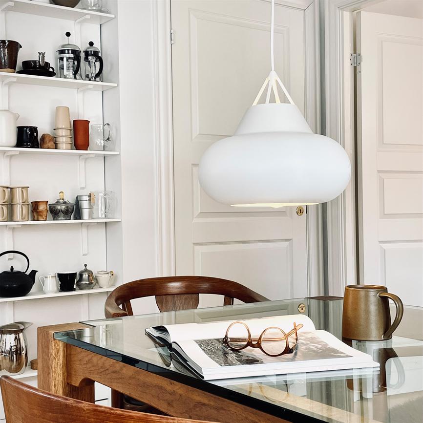A white ceiling lamp hangs above a glass table; on the table is an open book with round glasses and a ceramic jug; surrounded by wooden chairs, shelf with mugs and bright doors.