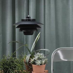Black tiered pendant lamp hangs above a group of succulents in terracotta pots on a table, in front of soft green curtains with a simple metal chair to the right.