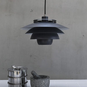 A dark gray layered pendant light hangs centrally, casting neutral light over a white tile table with a granite mortar and a stack of stainless steel containers; background: raw, gray concrete wall.