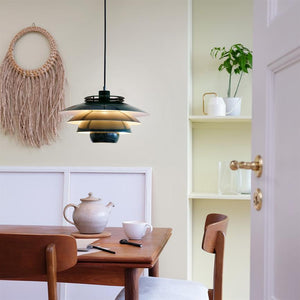 A layered black-gold pendant hangs over a wooden dining table; a teapot and mug are on the table, a chair pulls out, wall decorations and open shelves are seen in a bright, calm room.
