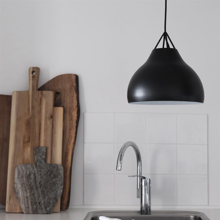Black pendant light hangs above a chrome kitchen faucet; in front of a white tile background, several wooden and stone cutting boards stand against the wall by the sink.