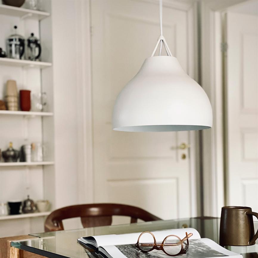 A white pendant lamp hangs above a glass table; an open magazine with brown glasses and a brown mug lie on the table, with a chair and shelf in a bright room.