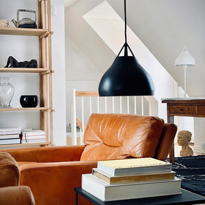 A cognac-coloured leather armchair sits under a large black pendant lamp, surrounded by a wooden bookcase and a side table with books in a bright loft with a pitched roof and staircase.