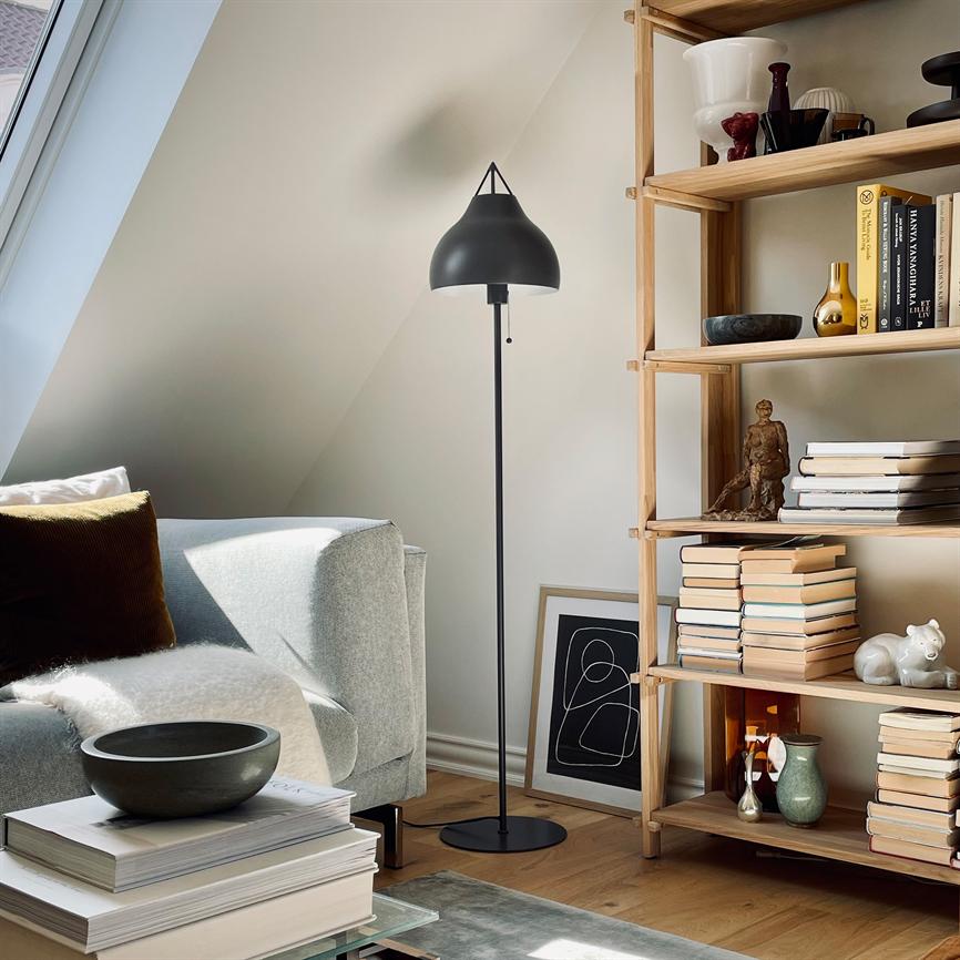 A floor lamp stands upright between a bright sofa and an open bookcase in a sunlit attic apartment, while books, bowls and a framed poster adorn the floor and shelves.