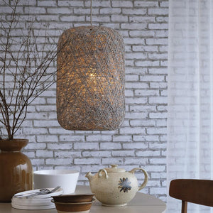 A wicker pendant lamp hangs and casts warm light over a table with a ceramic teapot, bowls and cutlery in front of a white brick wall and a light curtain.
