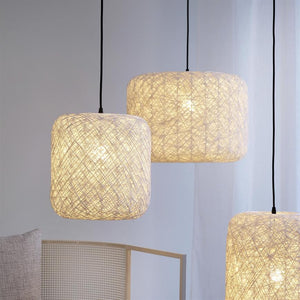 Three square lampshades made of twisted wire emit warm, diffused light, hanging in black joints above a bright room with a light curtain and a natural-coloured wicker chair.