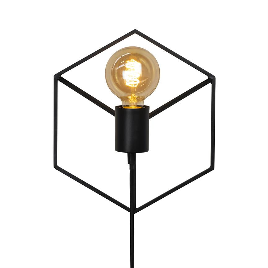 Black metal cube lamp radiates a warm, lit incandescent; the bulb sits centrally in an open, geometric frame, with a power cord hanging down against a white background.
