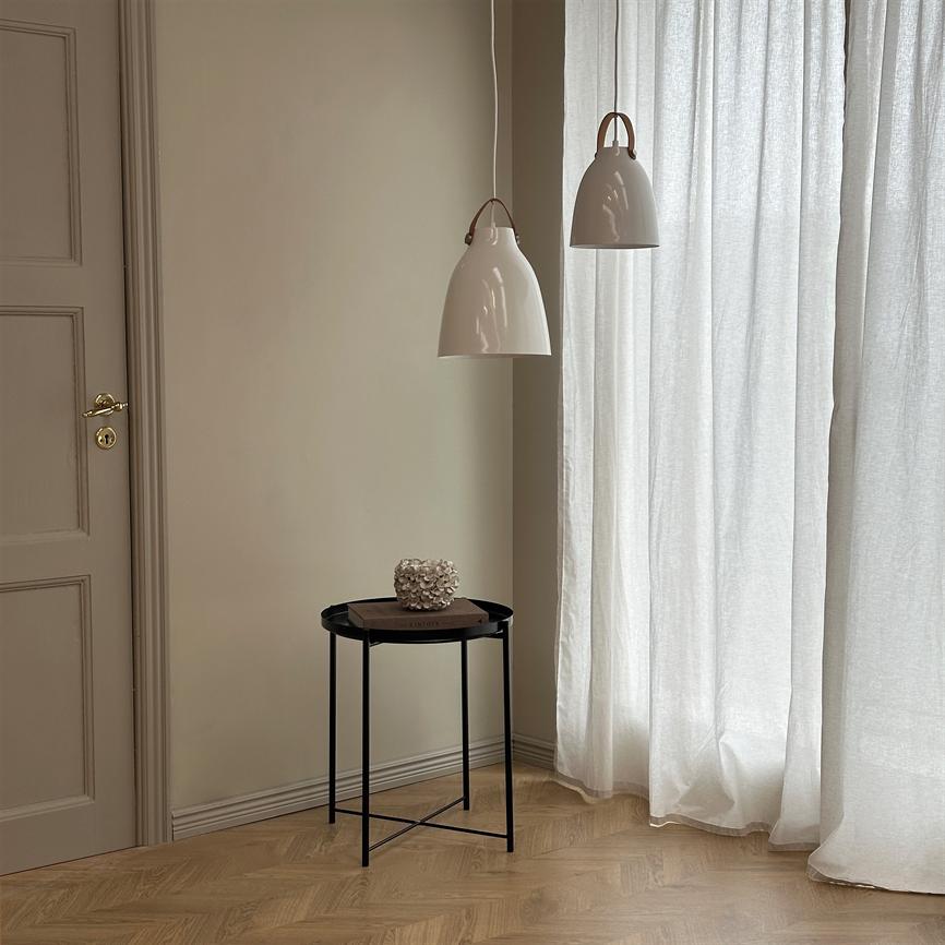 A small round black side table stands in a corner and carries a book and a white coral-like decoration under two white pendant lamps in front of slightly translucent curtains and a beige wall.