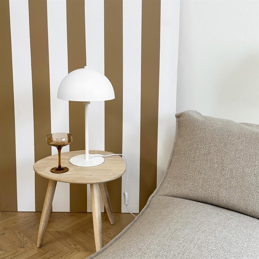 A small round wooden side table holds a white table lamp and a brown wine glass in front of vertical white and brown stripes, next to a beige sofa on a wooden floor.