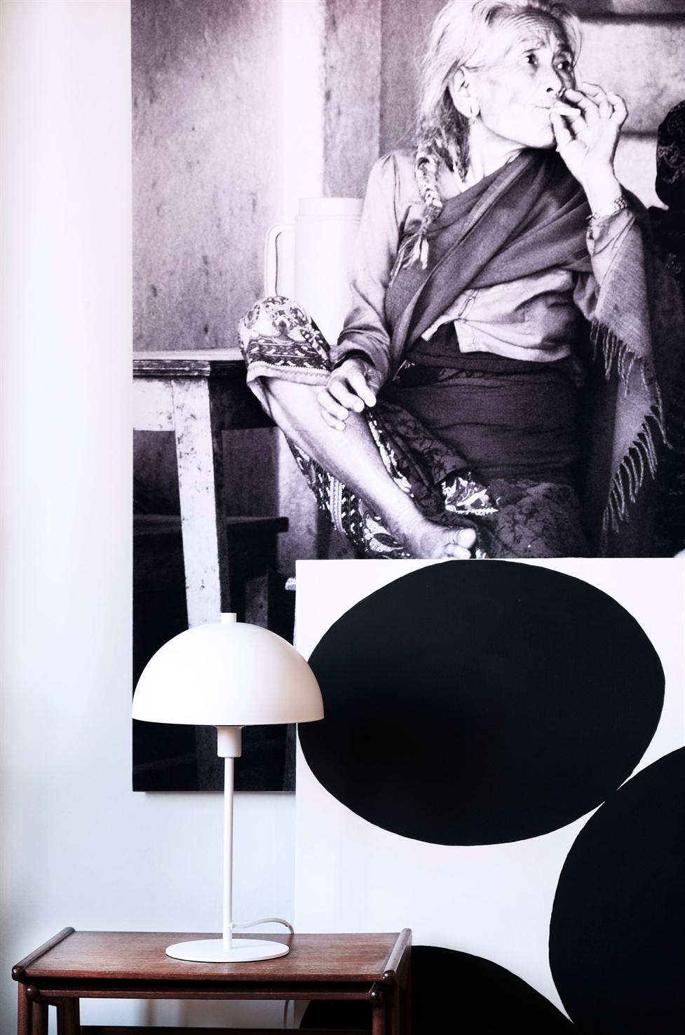 Lamp stands on a wooden table; in front of a large graphic canvas with black circles and a black and white photo of an elderly woman smoking in the background.