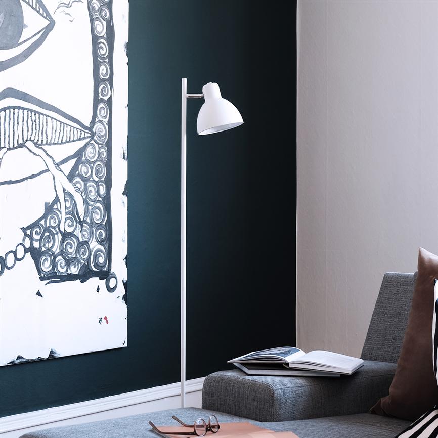 A white floor lamp stands by a gray sofa and points to a dark green wall; a black-and-white painting hangs on the left, book and glasses lie on the sofa.