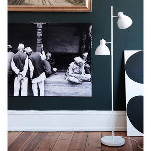 Large black-and-white photograph shows a group of elderly men in traditional hats, seated and conversing; hung on a dark green wall by a white floor lamp.