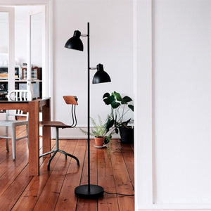 A black floor lamp stands upright by a wooden table and a metal-wooden chair; in the background potted plants, white walls and warm wooden floorboards in a bright, minimalist dining/work area.