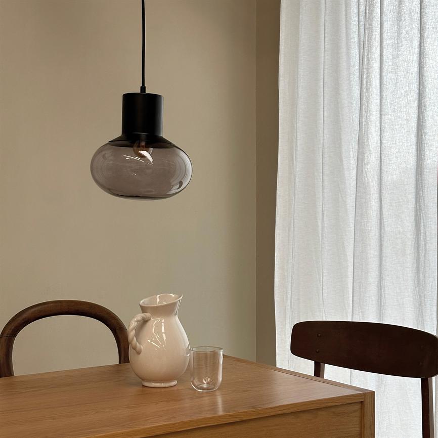 A dark glass pendant hangs over a wooden table; a white ceramic jug and a clear glass are on the table, with two wooden chairs and light, transparent curtains in the background.
