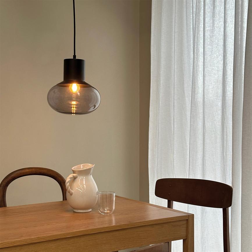 Hanging lamp shines with a warm glow above a wooden table; on the table is a white jug and a glass; the stage is surrounded by two wooden chairs and light, transparent curtains.