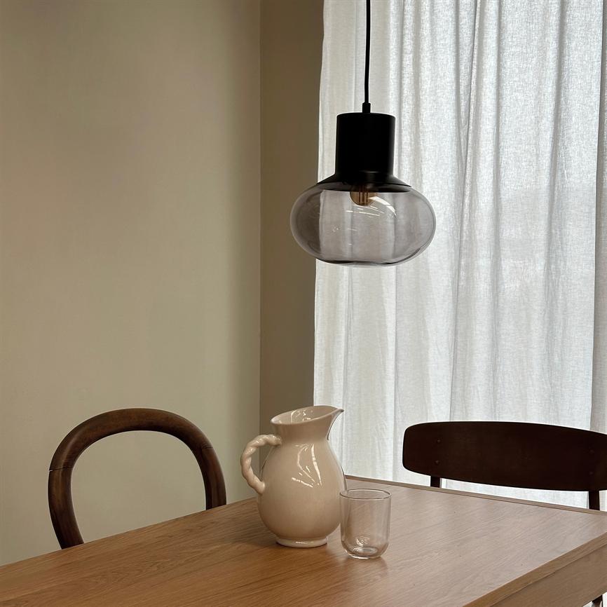 White ceramic jug stands on a wooden table by a clear glass; above them hangs a black pendant lamp with smoked glass, surrounded by two dark wooden chairs and a light-filtering curtain.