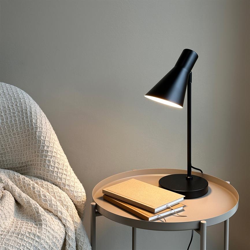 A black table lamp shines down on a round beige metal table, where two notebooks are stacked; next to it, a bright, textured rug sits over a chair against a neutral wall.