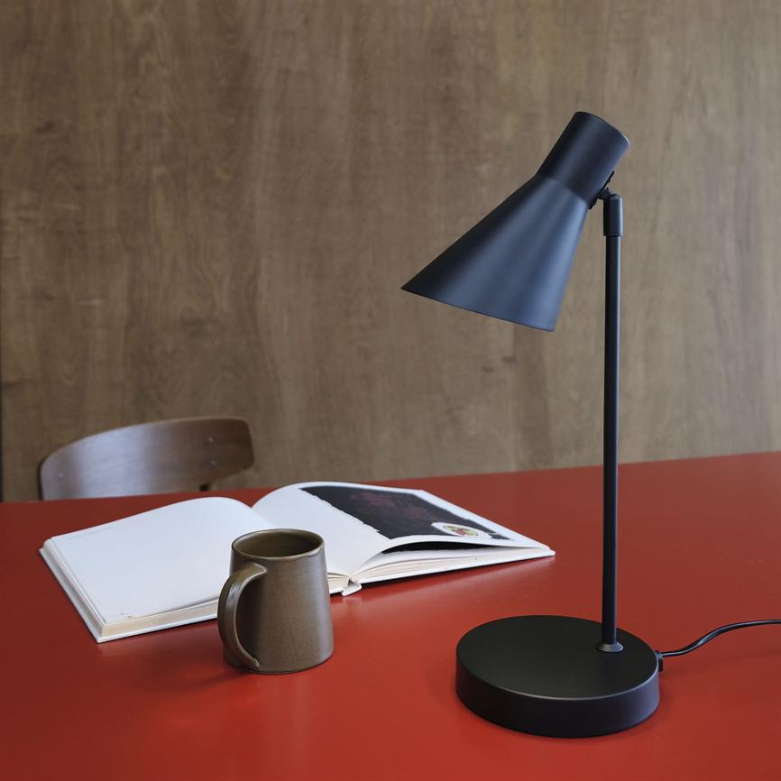 Black table lamp points down towards an open book and a brown mug on a red table, in front of a wooden wall and a wooden chair in the background.