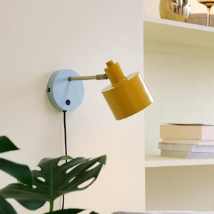 Yellow wall lamp with light blue round wall plate, pointing obliquely downwards; mounted on a cream-colored wall by a shelf with a stack of books, and a green monstera leaf in the foreground.