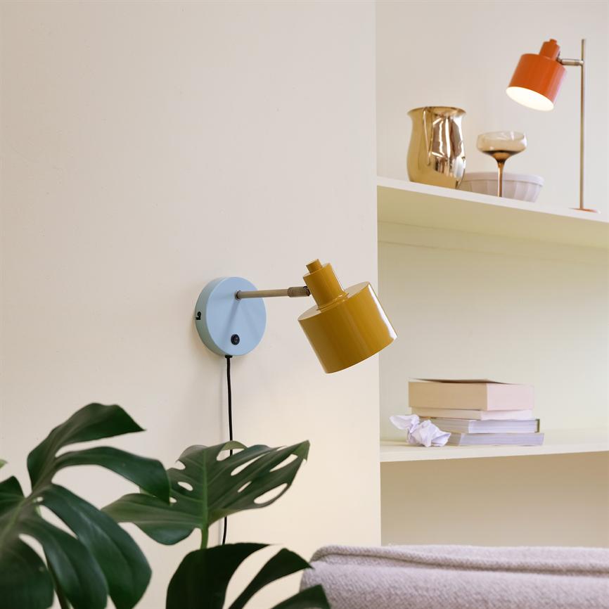 Yellow wall lamp sits on a light blue round plate and points obliquely downwards; the pale yellow wall is flanked by a shelf of books and decorations, and green plant leaves are seen in the foreground.
