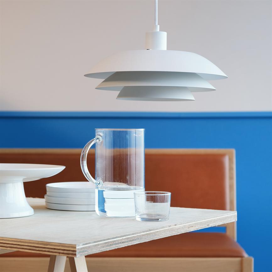 A white pendant lamp hangs above a light wooden table, where a clear glass jug, a glass, white plates and a footed bowl stand in front of a blue wall and brown leather bench.