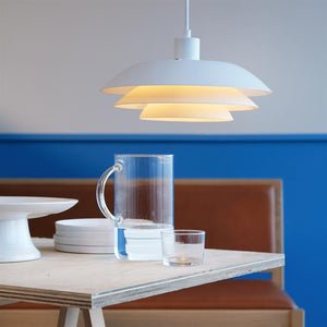 White layered hanging lamp emits warm light over a light wooden table with glass jug, glasses and stacks of white plates, in front of blue wall and brown upholstered bench.