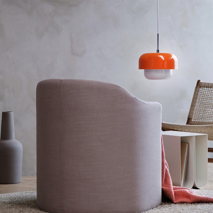 A pink upholstered armchair faces away from the viewer; a coral colored blanket falls down next to it. Minimalist living room with orange pendant lamp, wicker chair, ceramic vase and side table.