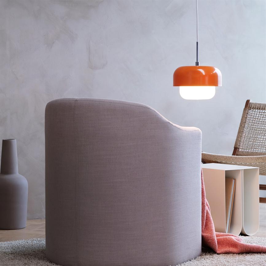 A pink upholstered armchair faces away, an orange hanging lamp shines above a low side table with books; a wicker chair, a rug and a vase stand in a calm, minimalist space.