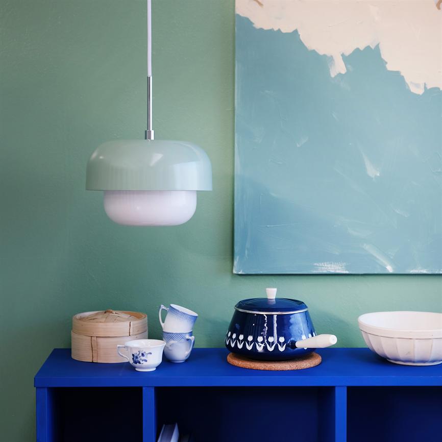 Lamp hangs above a blue cabinet; a blue patterned pot stands on the cabinet with cups, a steam basket and a white bowl in front of a green wall and abstract painting.