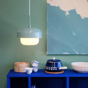 A suspended pastel green lamp shines above a cobalt blue chest of drawers, where teacups, a bamboo steam basket and a dark blue pot stand, in front of a green wall with an abstract painting.