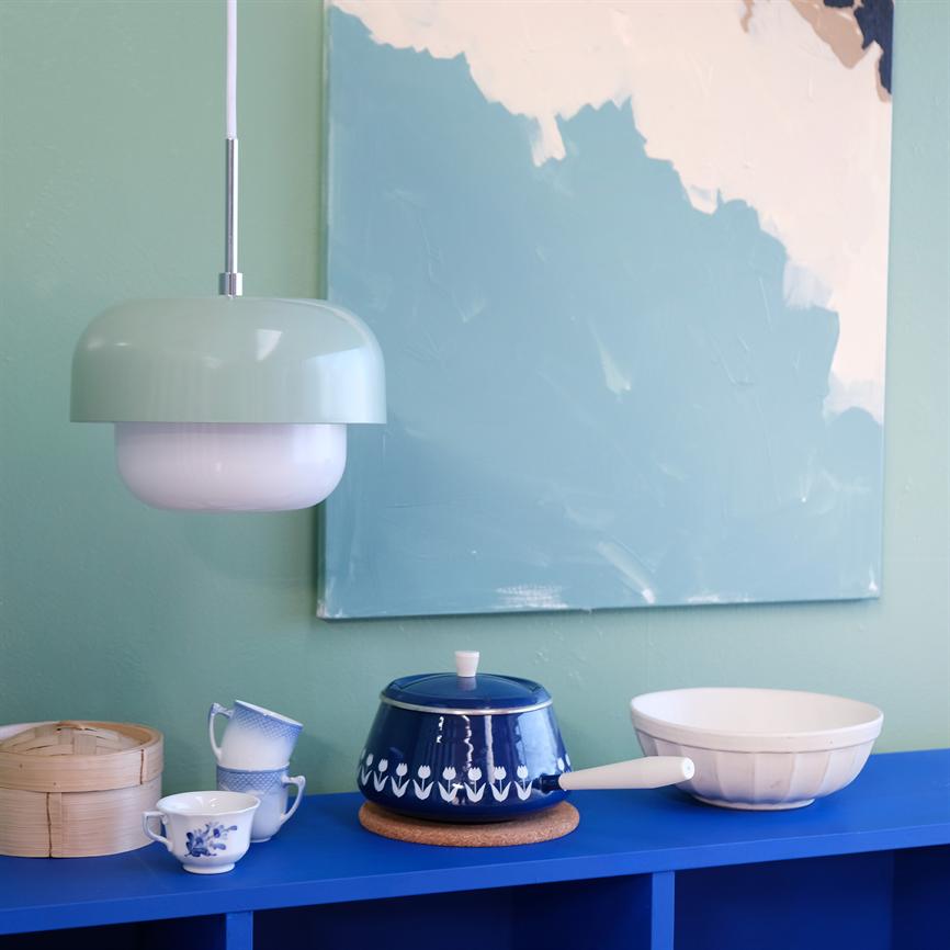 A mint green pendant light hangs above a blue bookcase; a blue pot with a lid stands on a cork base, surrounded by teacups, bamboo basket and a white bowl, with abstract blue painting behind.