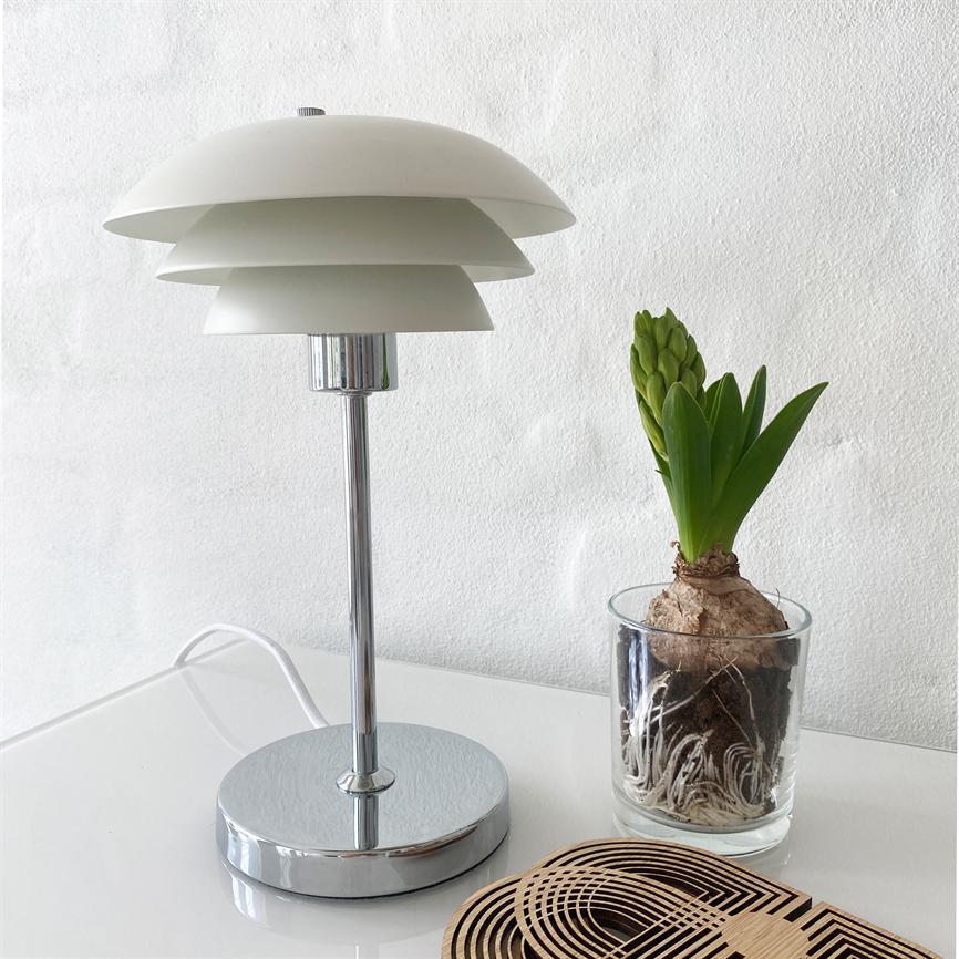 Table lamp with white, layered shade stands switched off on a white table; next to it stands a glass hyacinth with visible roots; white structural wall in the background.
