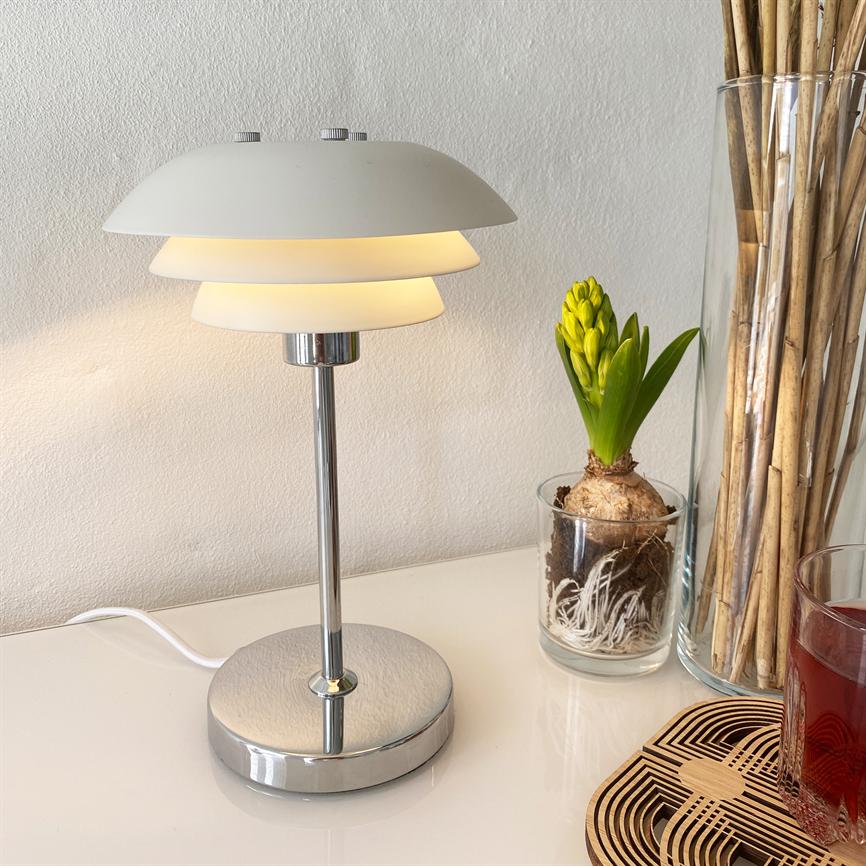 Table lamp emits warm, soft light; stands on a white tabletop by a light wall, by a glass hyacinth, a vase with dry branches and a glass of red drink on a wooden base.