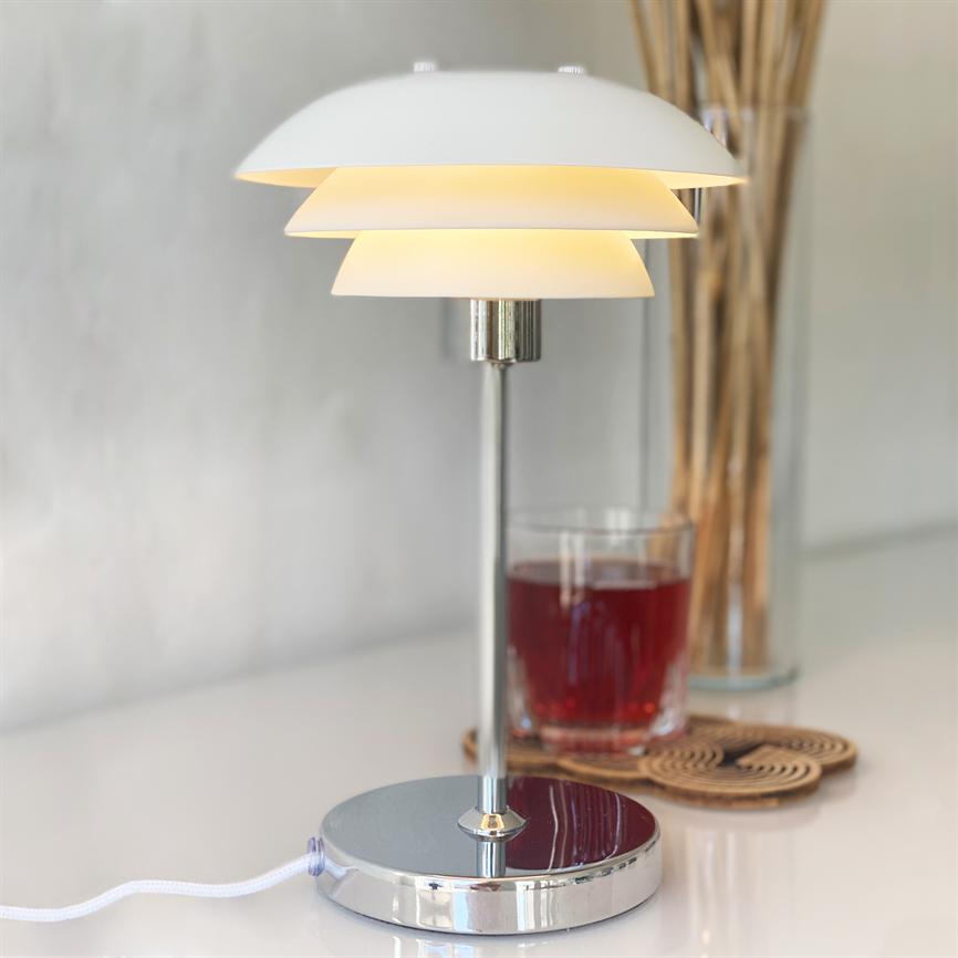 Table lamp with white, layered shade glows warm; stands on chrome feet on a white tabletop next to a glass of red drink and a tall glass vase with reeds.