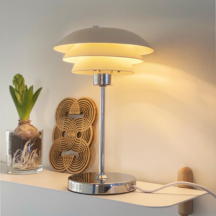 Chrome table lamp shines with layered white shades, casting soft warm light on a white wall, placed on a shelf by a glass plant with bulbs and a decorative wooden disc.