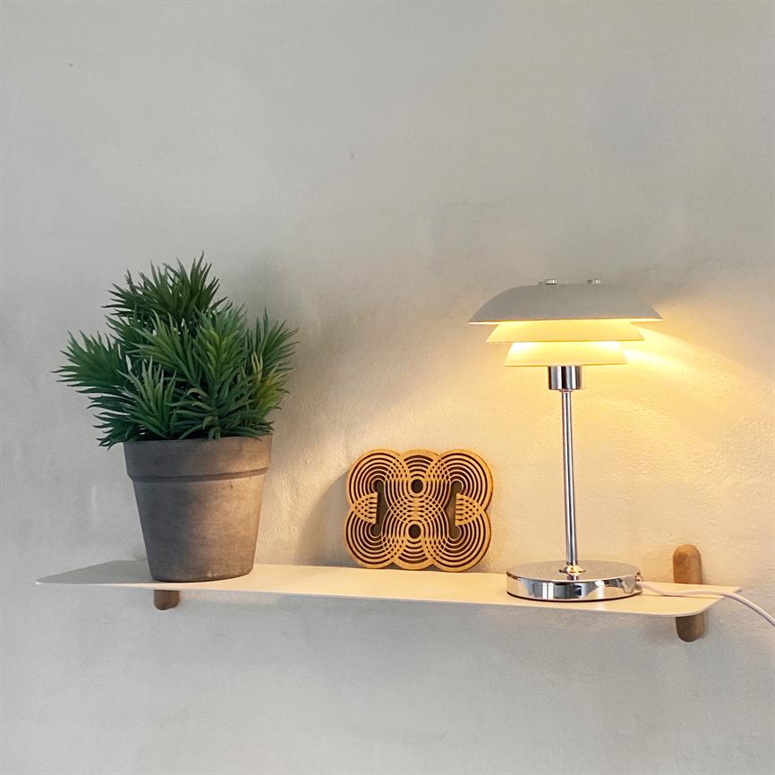Table lamp casts a warm, subdued glow over a white wall shelf; on the shelf stands a gray flowerpot with a green plant and a decorative wooden ornament against a light wall.