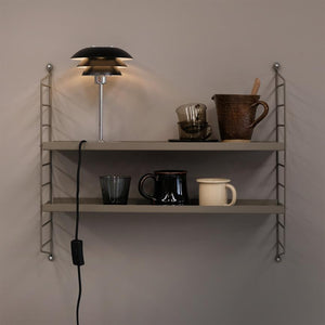 A wall-mounted two-shelf metal shelf holds a lit table lamp with a black shade, glass and ceramic cups, a rustic jug and a wooden spoon; cord with on/off button hangs down in front of a beige wall.