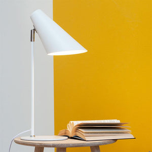 A white table lamp casts soft light over an open book on a round wooden table in front of a partition wall in white and mustard yellow.