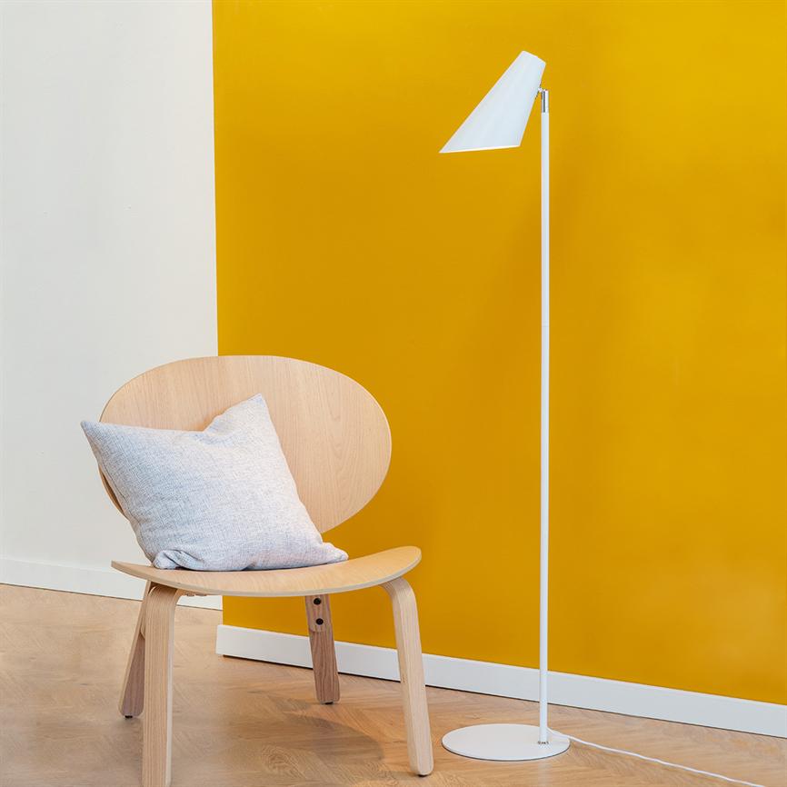 A light wooden chair stands with a light gray cushion, next to a white floor lamp in front of a deep yellow wall and light wooden floor.