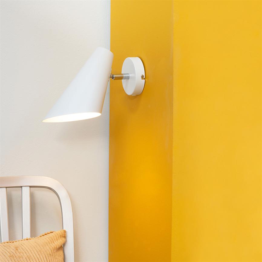 Wall lamp mounted on the wall shines downwards in a room with contrast between white and warm yellow wall, next to a white headboard with a yellow pillow.