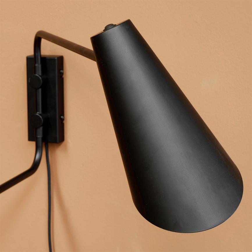 Black wall lamp with conical shade points downwards from a curved arm, mounted on a rectangular wall bracket; a black cable hangs down in front of a peach-colored wall.