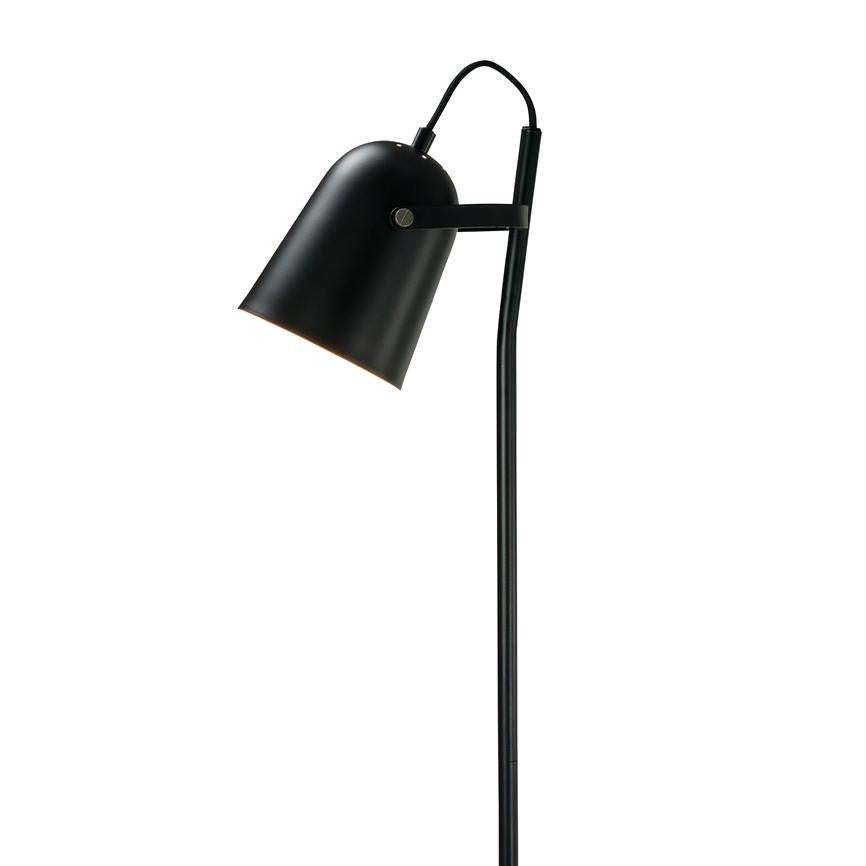 A black metal floor lamp leans slightly forward, the lampshade points downwards and emits soft light; slim stand against a uniform white background.