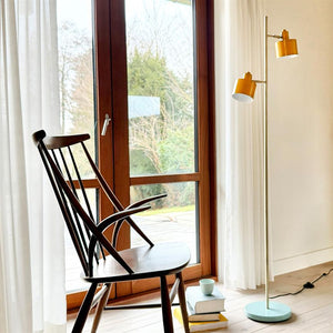 A standing lamp with yellow shades lights up next to a low light lounge chair; behind shelves with decorations (jug, glass, books) in a quiet, bright room seen through open white door.