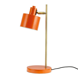 An orange metal table lamp tilts with its cylindrical shade downwards; it stands on a round orange base with a slender brass rod and black cord against a white background.